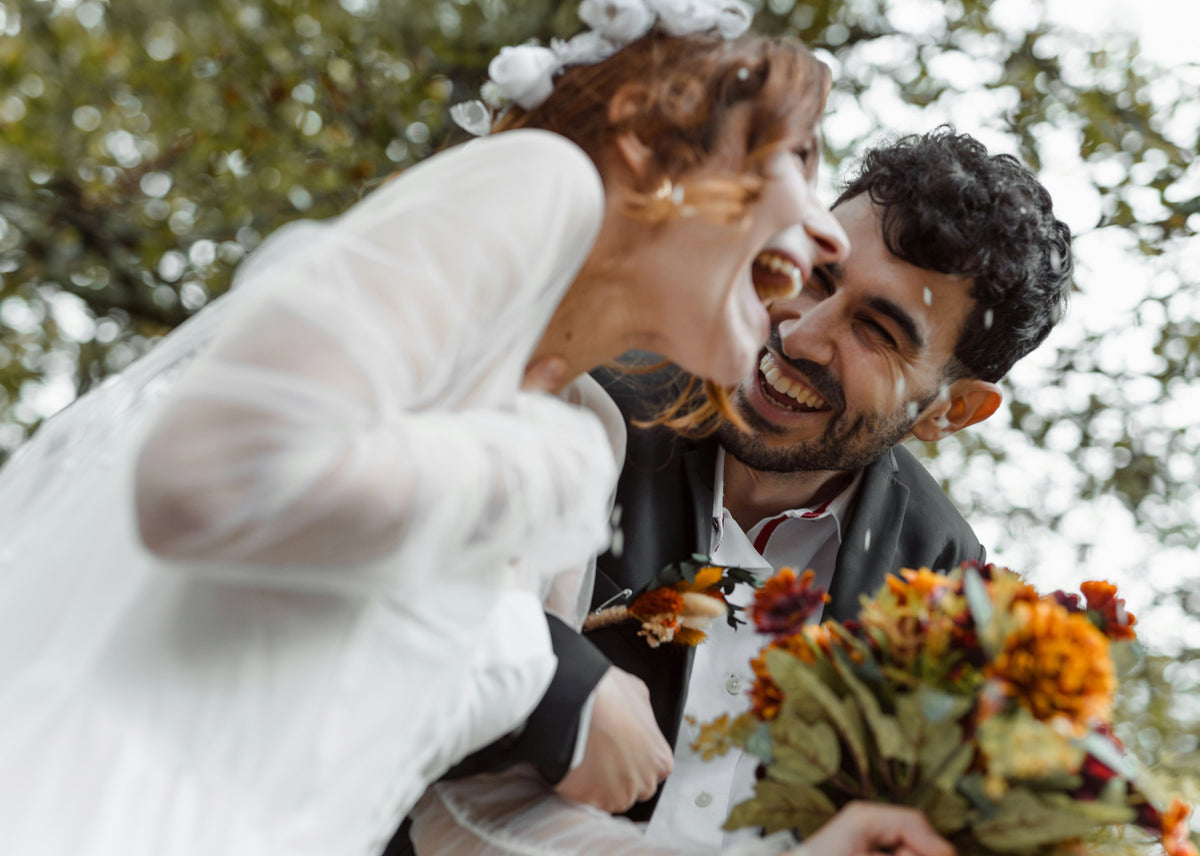A smiling bride in a white wedding dress leans in close to her groom outdoors, with confetti falling around them. The groom wears a dark suit jacket and white shirt, and the bride holds a bouquet of orange and burgundy flowers.