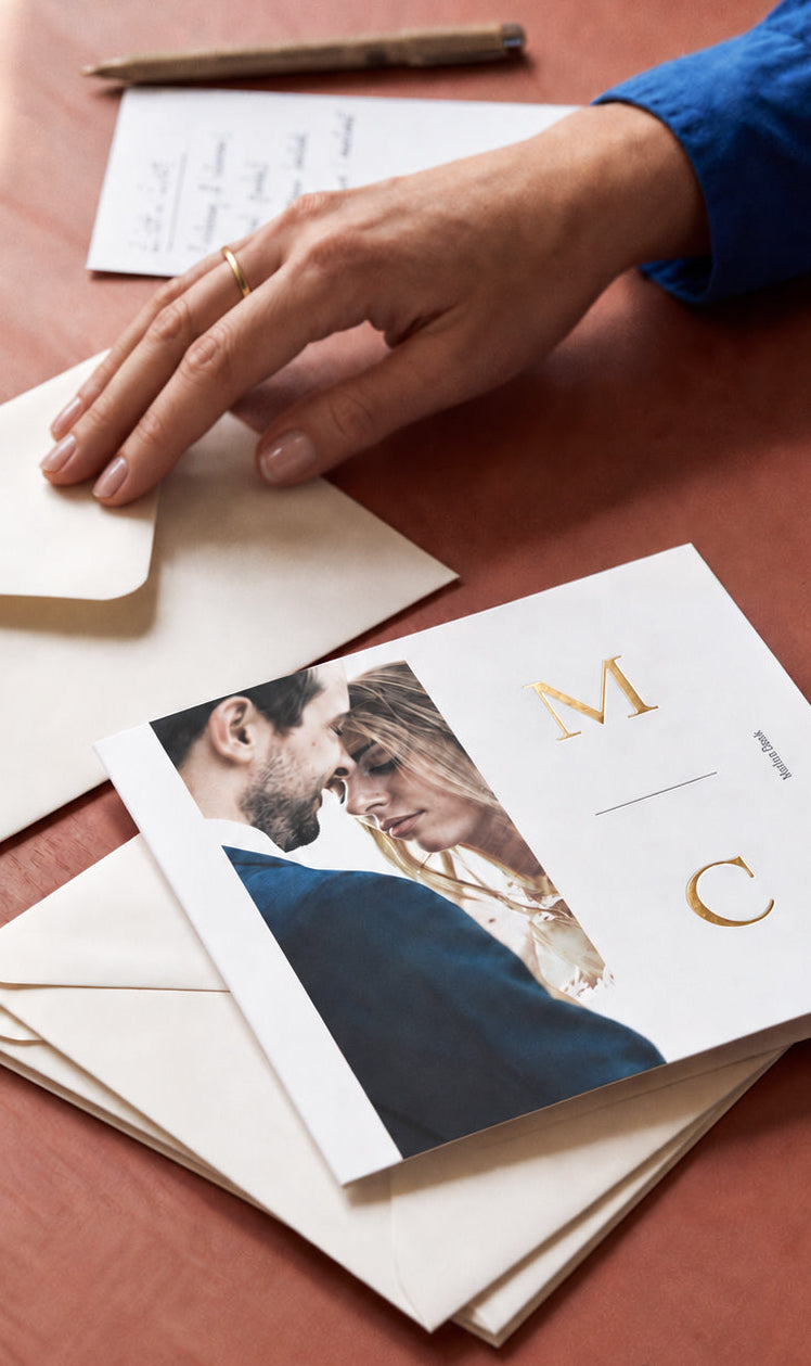 A styled flat-lay of a custom wedding invitation card and matching cream envelopes on a warm reddish-brown wooden table. The invitation features a photo of a couple and elegant gold monogram letters 'M' and 'C'.