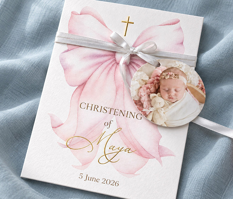 A christening invitation for Maya with a pink bow, a gold foil cross and a photo tag on a ribbon