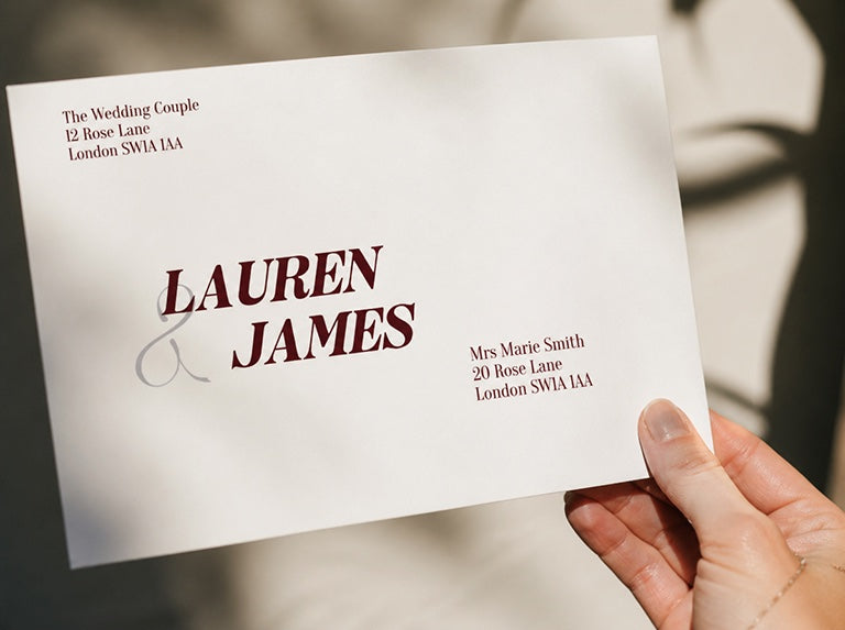 Hand holding an envelope with the couple's names in large burgundy letters and the sender and recipient addresses