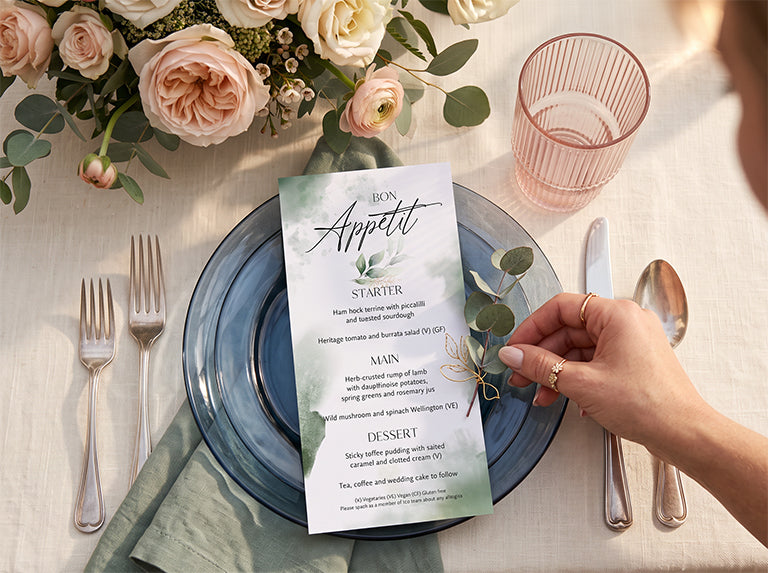 Green watercolour wedding menu card with a eucalyptus sprig on a blue glass plate, beside pink roses