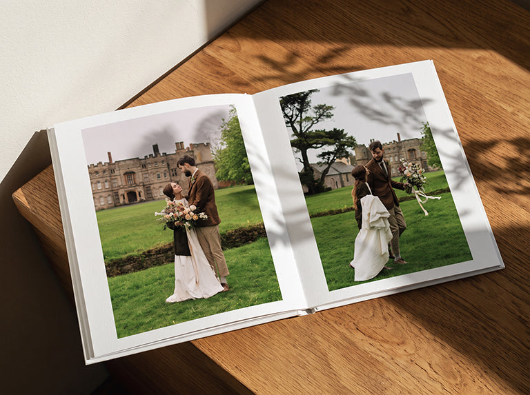 Open wedding photo book lying flat on a wooden surface, with photos of a bride and groom outside a country house