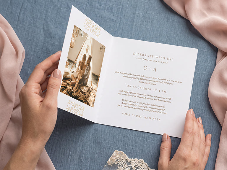 Hands holding an open folded card with a couple's photo and gold lettering, on a blue linen cloth
