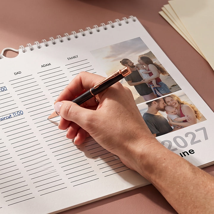 A hand writing in a family calendar with a column for each person, beside family photos